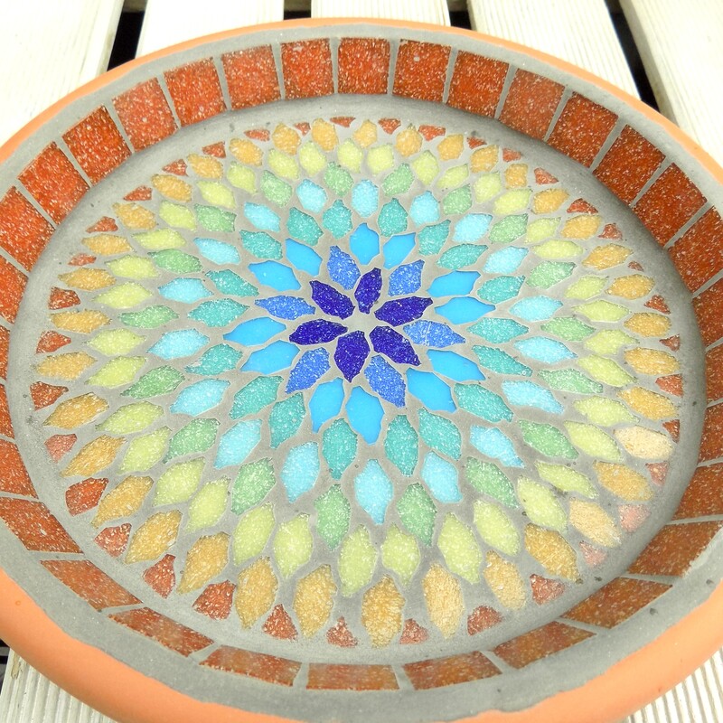 A mosaic bird bath with a  mandala style design in the colours of the rainbow from terracotta on the outside to indigo blue in the centre