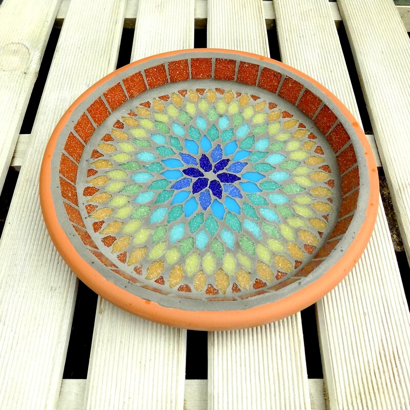 A mosaic garden bird bath with a  mandala style design in the colours of the rainbow from terracotta on the outside to indigo blue in the centre